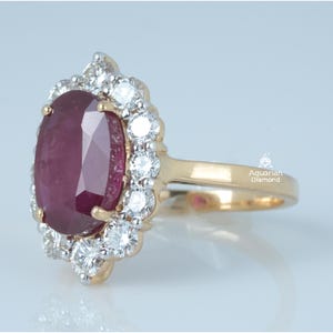 Könnte beinhalten: Ein Goldring mit einem großen, oval geschliffenen, tiefroten Edelstein, der von einem Kranz aus kleinen, runden, weißen Steinen umgeben ist. Die Ringschiene ist aus poliertem Gold und der Schriftzug Aquarian Diamond ist seitlich zu sehen.