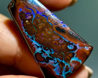 May include: A polished boulder opal gemstone with a trapezoidal shape, featuring a dark brown ironstone matrix contrasted by vibrant, iridescent veins of electric blue and purple play-of-color that shimmer across the smooth, reflective surface.