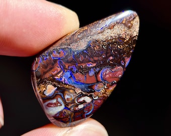 May include: A polished boulder opal gemstone with a teardrop shape, featuring a dark brown ironstone matrix with vibrant flashes of electric blue, deep red, and metallic gold colors swirling throughout the surface in a natural pattern.