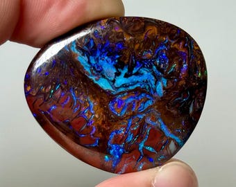 May include: A polished boulder opal gemstone with a dark brown ironstone matrix featuring vibrant, iridescent flashes of electric blue, purple, and green light throughout the surface in a swirling, organic pattern with a smooth, rounded shape.