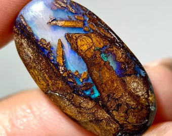 May include: An oval-shaped boulder opal gemstone featuring a polished surface with deep brown ironstone matrix veins interspersed with vibrant flashes of iridescent blue and green color play throughout the stone's smooth, rounded face.