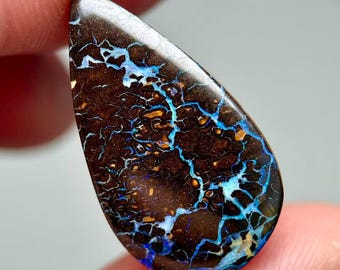May include: A teardrop-shaped boulder opal gemstone featuring a dark brown matrix base with intricate, branching veins of vibrant electric blue and subtle golden-orange flashes throughout the polished, smooth surface of the stone.