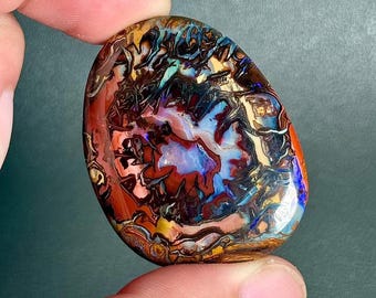 May include: A polished, oval-shaped boulder opal gemstone featuring a complex, swirling pattern of deep red, burnt orange, dark brown, and vibrant electric blue colors. The stone has a smooth, glossy surface with a high-contrast, multi-colored play of light.