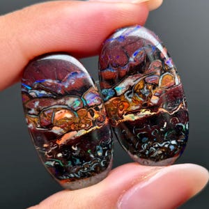 Puede incluir: Dos ópalos boulder de forma ovalada con una matriz de piedra de hierro marrón oscuro y destellos vibrantes de juego de colores iridiscentes en azul, verde y naranja. Las gemas pulidas muestran patrones naturales complejos y una superficie lisa.
