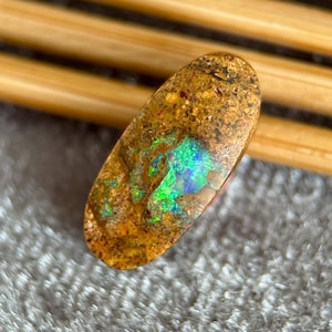 May include: An oval-shaped opal gemstone with a brown matrix and vibrant flashes of green and blue. The polished stone displays a play of color, showcasing a spectrum of hues. The opal is set against a neutral background.