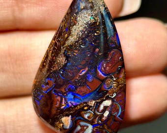 May include: A teardrop-shaped boulder opal gemstone featuring a dark brown ironstone matrix with vibrant flashes of electric blue, deep purple, and metallic gold color play throughout the polished, smooth, and reflective surface of the stone.