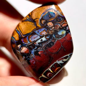May include: A polished boulder opal gemstone featuring a complex matrix of deep red, earthy brown, and dark charcoal tones. The surface displays vibrant flashes of iridescent blue and violet light, with intricate natural veining throughout.