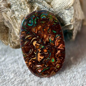 May include: An oval-shaped boulder opal gemstone featuring a dark brown ironstone matrix with swirling patterns of iridescent green, blue, and orange flashes. The polished surface displays a high-gloss finish with intricate, organic color veins.