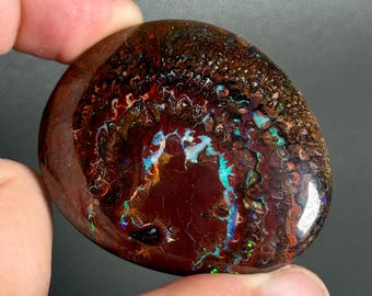 May include: A polished boulder opal gemstone with a dark brown ironstone matrix featuring vibrant flashes of iridescent blue, green, and orange color play across its smooth, rounded surface, showcasing a natural, intricate, and organic pattern.