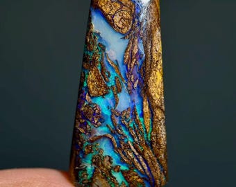 May include: A polished boulder opal gemstone with a trapezoidal shape, featuring a complex matrix of iridescent blue and green flashes embedded within a textured, earthy brown ironstone host rock with a smooth, reflective surface finish.