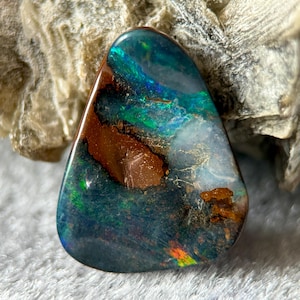 Puede incluir: Una piedra preciosa de ópalo boulder triangular pulida que presenta una base de matriz de piedra de hierro con destellos vibrantes de colores iridiscentes verdes, azules y naranjas en su superficie lisa y reflectante, mostrando inclusiones minerales naturales.