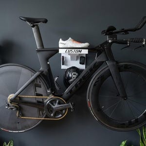 Peut inclure: Un vélo de route noir monté sur un mur, avec une chaussure blanche et des accessoires sur une étagère avec l'inscription "CUSTOM". Le vélo a des roues noires, un cadre noir et une chaîne dorée. Des plantes vertes encadrent le vélo.