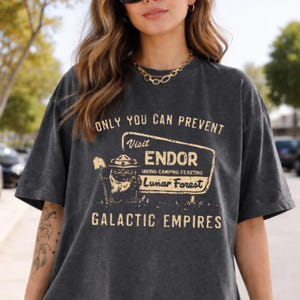 May include: Charcoal grey t-shirt featuring the text "ONLY YOU CAN PREVENT" above a sign that reads "Visit Endor Hiking-Camping-Feasting Lunar Forest Galactic Empires". The shirt has a faded, vintage appearance.