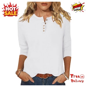 May include: A white long-sleeve henley shirt featuring a round neckline with a vertical placket containing five small brown buttons. The shirt is worn tucked into blue denim jeans secured with a brown leather belt with a gold-tone buckle.