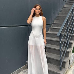 May include: A white sleeveless dress with a high neckline and a sheer, flowing skirt. The dress has a fitted bodice and a long, elegant skirt. The model is standing on a set of outdoor stairs.