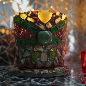 Könnte beinhalten: Ein Mosaik-Kerzenhalter mit herzförmigem Akzent. Der Halter ist zylindrisch und besteht aus farbigen Glasstücken in Grün-, Rot-, Gelb- und Grautönen. Eine kleine rote Kerze ist in der Nähe angezündet.