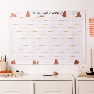 May include: A white 2026 year planner featuring bear illustrations. The calendar has a grid layout with horizontal lines and bear graphics. The text "2026 YEAR PLANNER" is printed at the top. The planner is on a white wall.