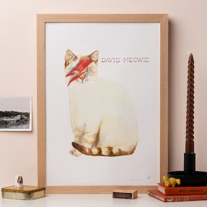 Op de afbeelding: Een aquarel schilderij van een witte kat met een rode en blauwe bliksemflits over zijn oog. De kat zit en kijkt naar rechts. De tekst "DAVID MEOWIE" staat in het zwart boven de kat.
