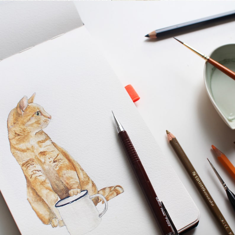 May include: A watercolor painting of an orange tabby cat sitting with a white mug. The cat is looking to the right of the image. The painting is on a white sheet of paper with a pencil and other art supplies on the table.