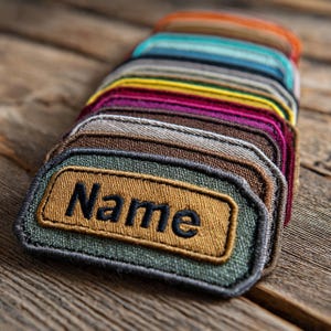 Op de afbeelding: Een verzameling rechthoekige patches in verschillende kleuren, elk met een contrasterende rand. De patches hebben een bruin rechthoek met het woord "Name" in zwart geborduurd. Ze zijn gerangschikt op een houten oppervlak.