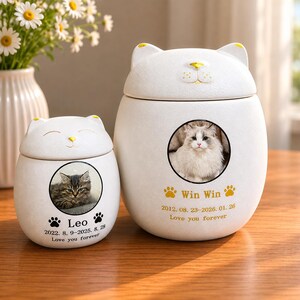 May include: Two white ceramic cat-shaped urns. The urns have a circular photo of a cat and text that reads "Leo" and "Win Win" with dates and "Love you forever". The urns have a lid with cat ears and a yellow nose.