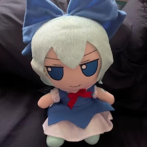 May include: A plush doll featuring a character with light blue hair and a large blue bow. The doll has blue eyes, a blue dress, a white skirt, and a red bow. The doll is standing on a dark surface.