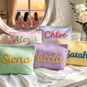 Puede incluir: Varias bolsas de cosméticos personalizadas en tonos pastel, como rosa, azul, verde, morado y amarillo. Cada bolsa tiene un nombre bordado en un color contrastante, hecha de tela seersucker a rayas.