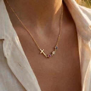May include: A gold-colored necklace with a cross pendant and three gemstone accents. The necklace is worn against a white shirt, with a blurred background of a field.
