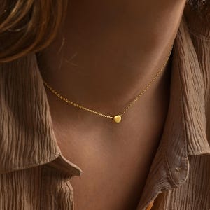 Pode incluir: Um delicado colar de ouro com um pequeno pingente em forma de coração. O colar tem um design de corrente simples, com o pingente de coração centrado. A cor dourada do colar contrasta com o tom de pele da pessoa que o usa.