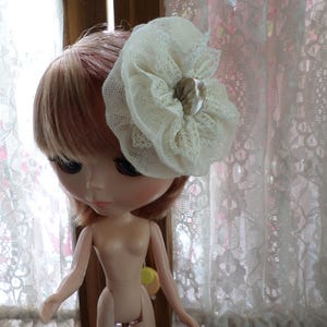 May include: A doll with long, pink hair wearing a white lace flower hair accessory with a pearl-like center.