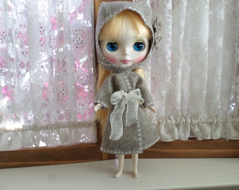 May include: A doll with blonde hair wearing a grey hooded coat with a white ribbon. The doll has blue eyes and is wearing a grey dress.