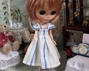 May include: A doll with auburn hair wearing a white dress with blue vertical stripes. The doll is standing on a white stand. The background includes miniature furniture and decor.