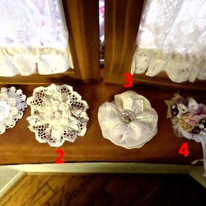May include: Four decorative fabric flower embellishments labeled one through four. Item one is an elongated white lace piece. Item two is a round white lace flower. Item three is a layered white fabric flower. Item four features fabric petals and beads.