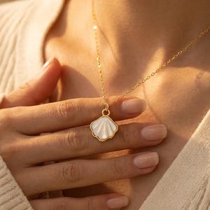 May include: A gold necklace featuring a seashell pendant. The pendant has a white shell design with a gold border. The necklace is being held by a person wearing a cream-coloured jumper.