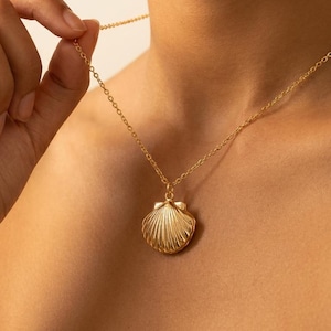 May include: A gold-colored necklace with a seashell pendant. The pendant is intricately detailed with ridged lines, and the chain is delicate. The necklace is worn around the neck, and the background is a blurred skin tone.