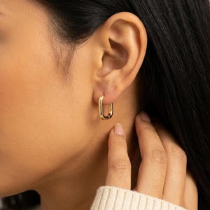 May include: Gold-colored rectangular hoop earrings. The earrings are worn on the ear of a person with dark hair. The earrings have a polished finish and a simple, modern design. The person is wearing a cream-colored sweater.