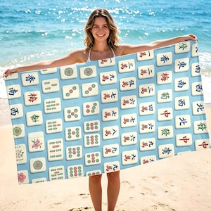 May include: A rectangular beach towel featuring a light blue background patterned with a grid of various mahjong tiles. The tiles display traditional Chinese characters, circular dot patterns, bamboo sticks, and floral designs in red, green, and black ink.