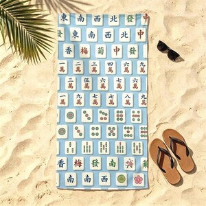 May include: A rectangular beach towel featuring a light blue background with a repeating pattern of white Mahjong tiles. The tiles display various traditional Chinese characters, numbers, and floral symbols in red, green, and black ink colors.