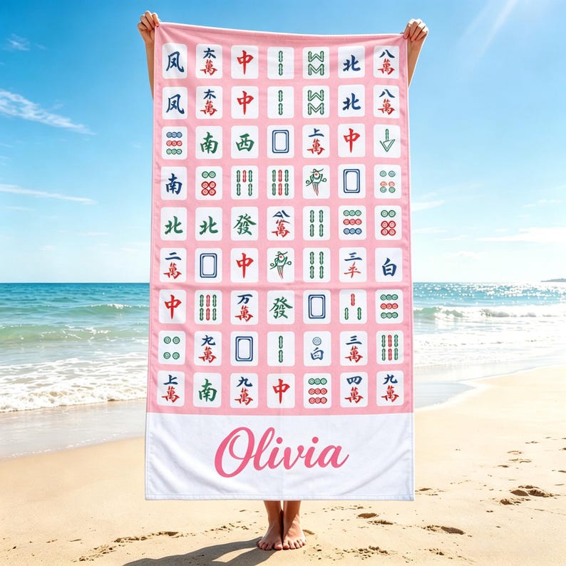 May include: A pink beach towel featuring a pattern of Mahjong tiles in various colors. The towel has a white border at the bottom with the name "Olivia" in pink script. The towel is held up against a sunny beach backdrop.