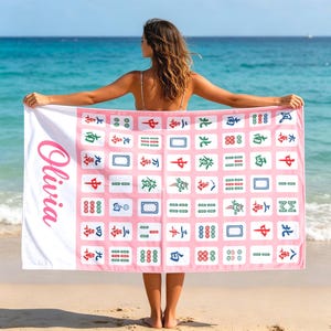 May include: A personalized beach towel with a pink and white design. The towel features a Mahjong tile pattern and the name "Olivia" in pink script. The background is a beach with blue water.