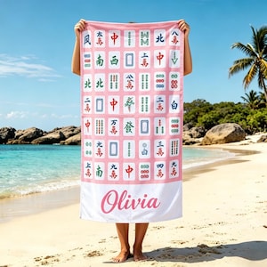May include: Pink beach towel featuring a Mahjong tile pattern with Chinese characters and symbols. The bottom section has the name "Olivia" in a cursive font. The towel is held up on a sandy beach with a blue sky and ocean in the background.