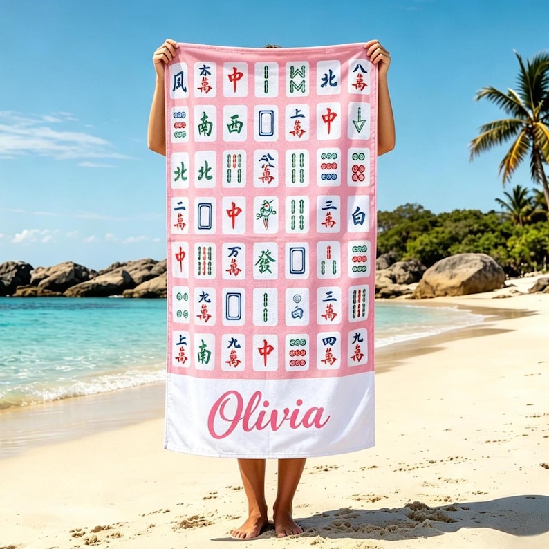 May include: Pink beach towel featuring a Mahjong tile pattern with Chinese characters and symbols. The bottom section has the name "Olivia" in a cursive font. The towel is held up on a sandy beach with a blue sky and ocean in the background.