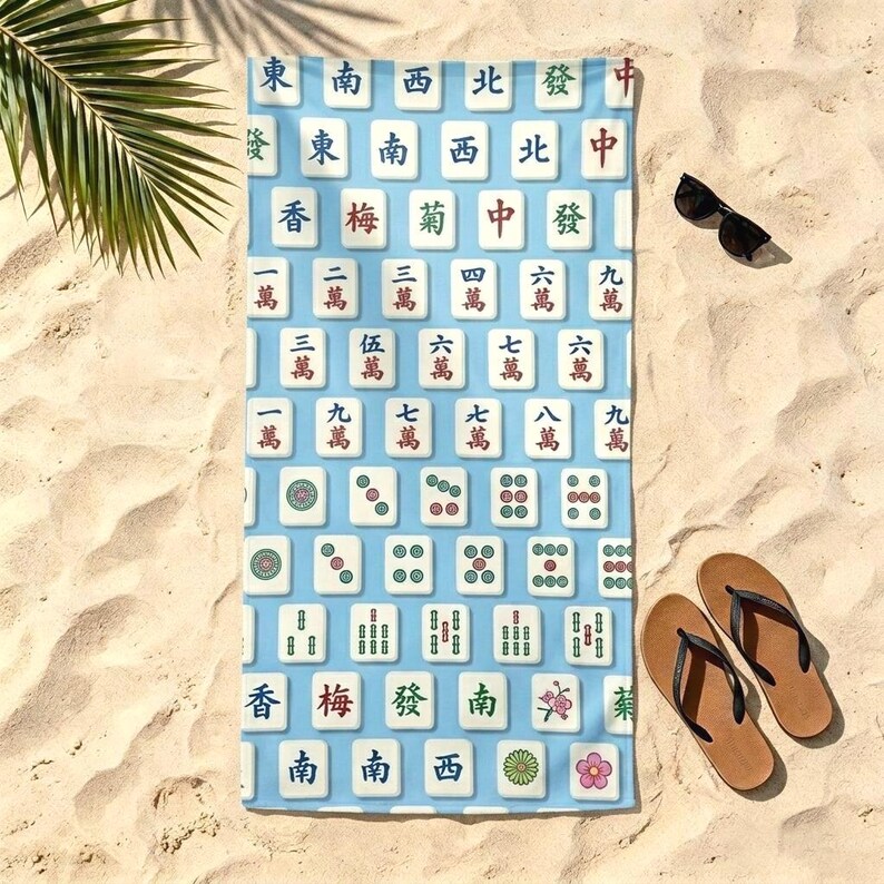 May include: A rectangular beach towel featuring a repeating pattern of white mahjong tiles on a light blue background. The tiles display various Chinese characters, numbers, and floral symbols in red, green, and black ink across the entire surface.