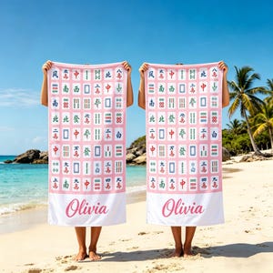 May include: Two pink beach towels with a Mahjong tile pattern and the name "Olivia" in cursive. The towels are held up on a beach with the ocean and palm trees in the background.