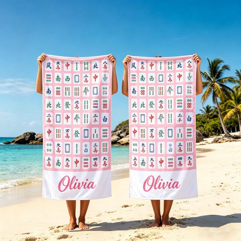 May include: Two pink beach towels with a Mahjong tile pattern and the name "Olivia" in cursive. The towels are held up on a beach with the ocean and palm trees in the background.