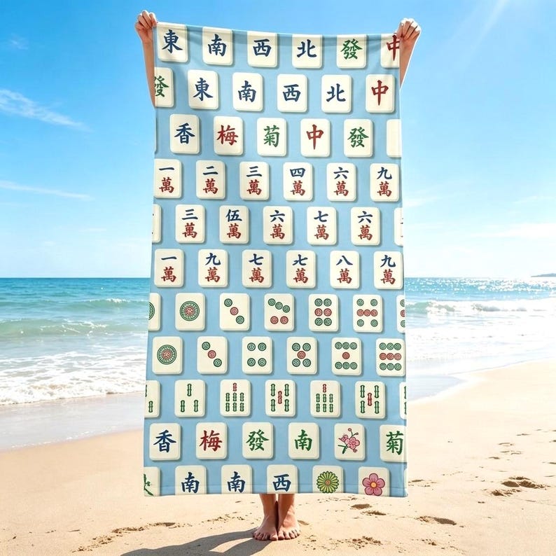 May include: A rectangular beach towel featuring a light blue background patterned with a grid of various Mahjong tiles. The tiles display traditional Chinese characters, numbers, and floral symbols in red, green, and black ink on a white base.