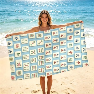 May include: A rectangular beach towel featuring a light blue background patterned with various mahjong tiles. The tiles display traditional Chinese characters, numbers, and floral symbols in red, green, and black ink, arranged in a repeating grid layout.