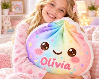 May include: A plush, dumpling-shaped pillow featuring a rainbow gradient design with a smiling face, pink cheeks, and small colorful stars. The name Olivia is printed in large, bold pink letters across the front of the soft, rounded cushion.