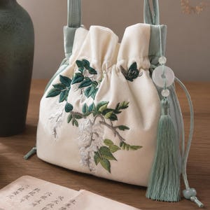 Könnte beinhalten: Cremefarbener Kordelzugbeutel mit gestickten grünen Blättern und weißen Blüten. Die Tasche hat eine hellgrüne Einfassung und einen Griff, mit einem passenden Quasten- und jadeartigen Perlenanhänger. Die Tasche liegt auf einer Holzoberfläche.