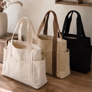 Può includere: Tre borse tote in tela disposte in fila, che includono una borsa color crema, una borsa beige con manici marroni e una borsa nera. Ogni borsa presenta due manici superiori, una tasca frontale con bottone a pressione dorato e due tasche laterali.